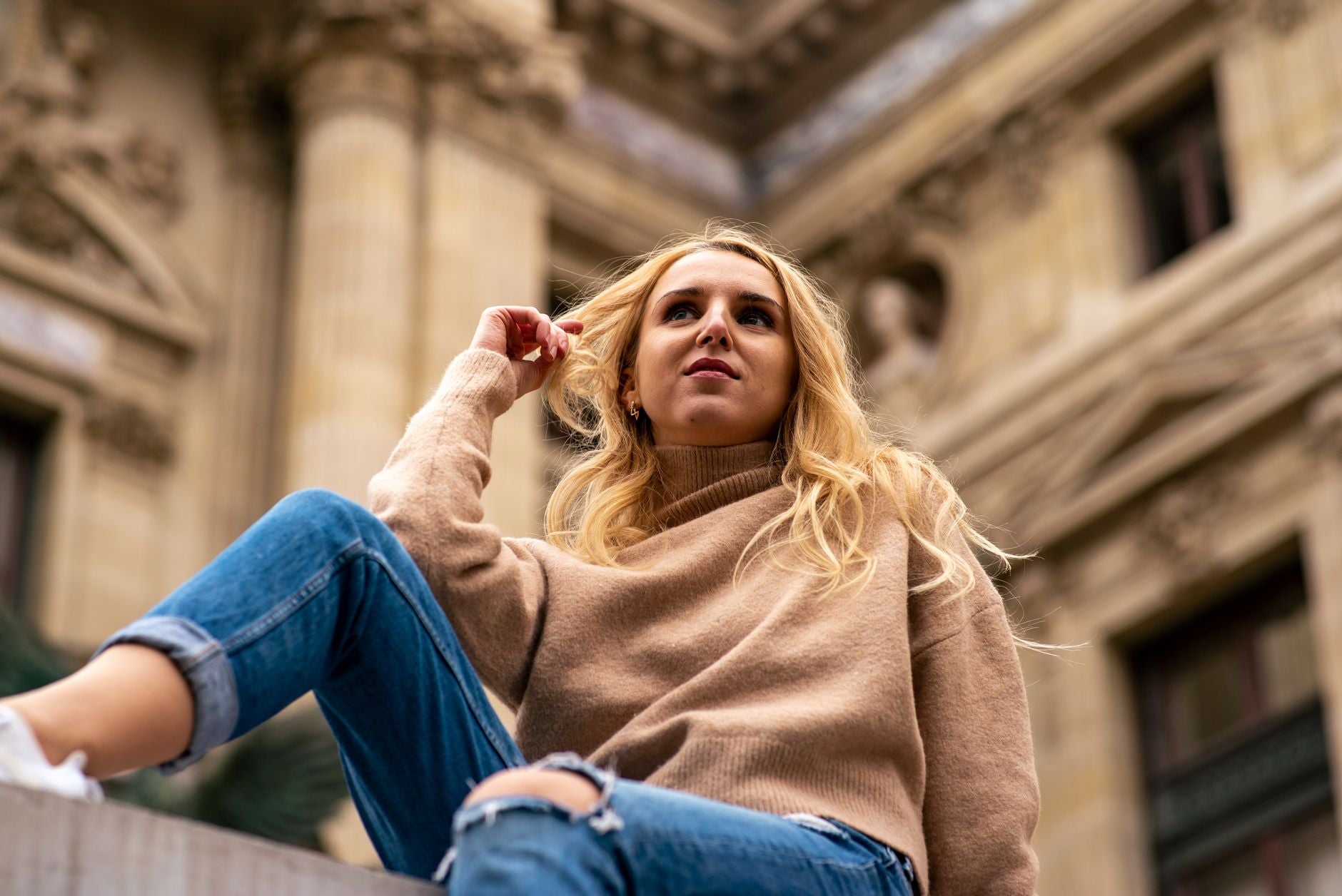 Femme en pull col roulé cachemire beige assise devant une façade haussmannienne
