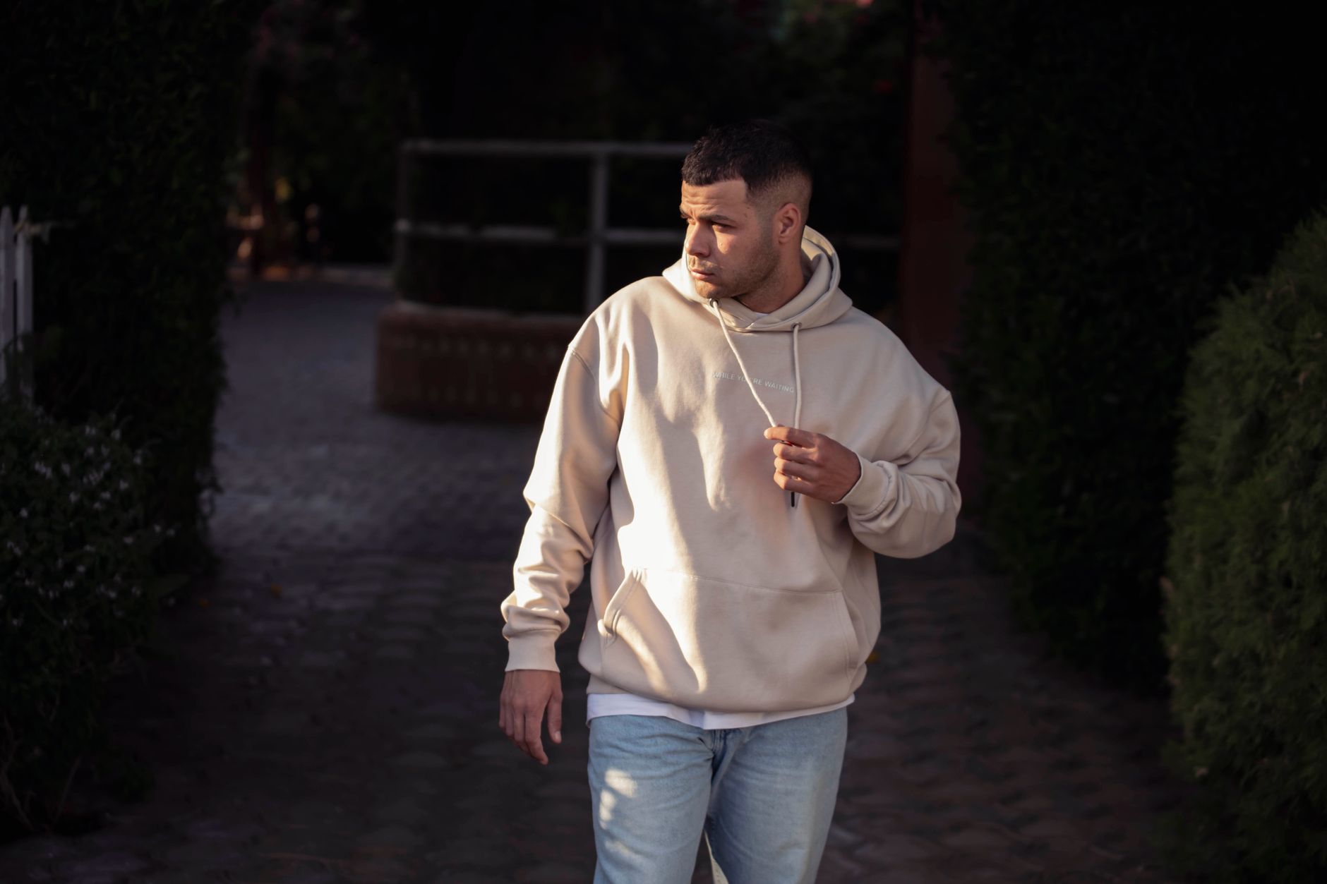 Homme en sweat à capuche beige oversize marchant dans une rue pavée urbaine